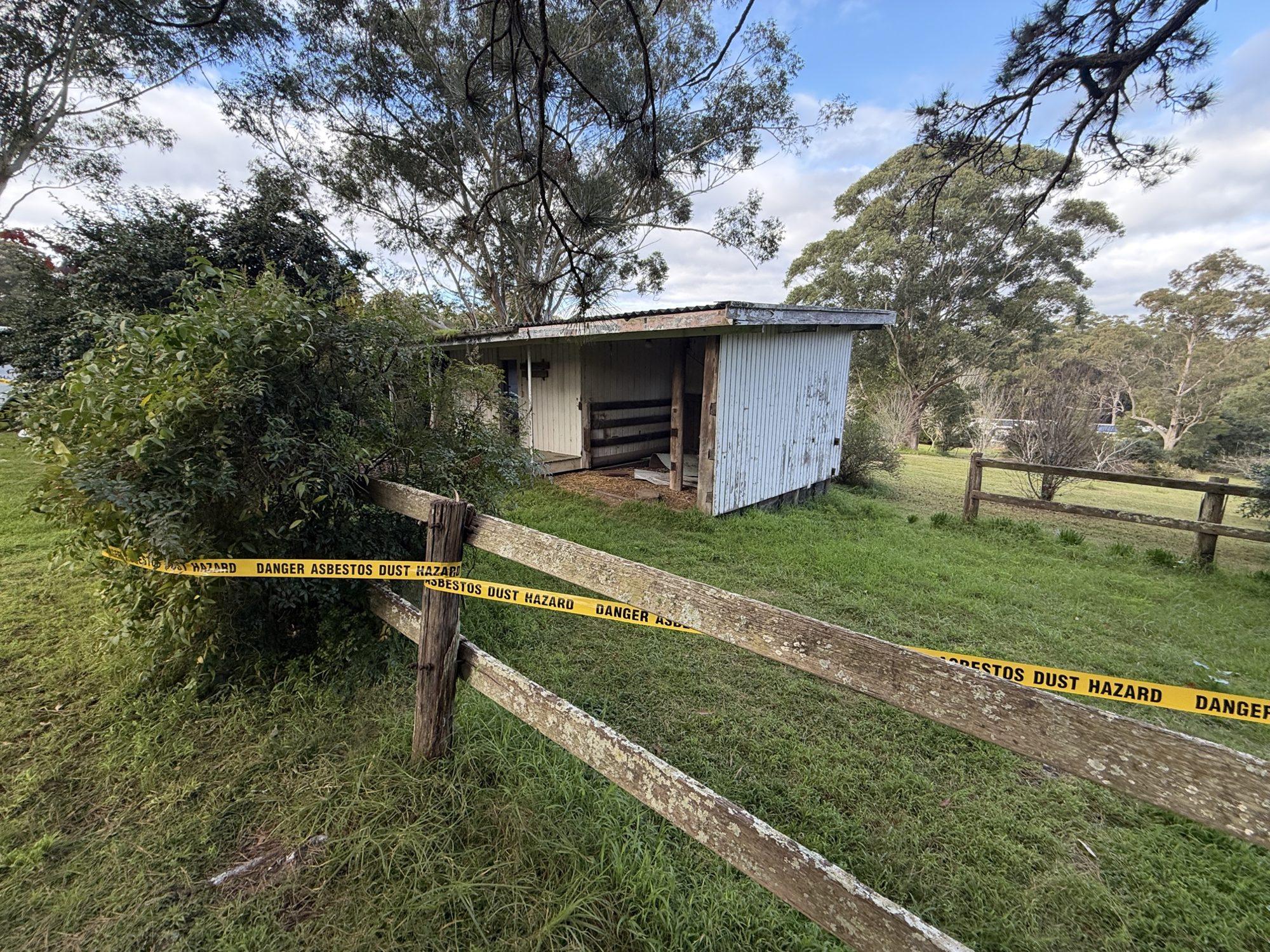 Yellow asbestos dust hazard tape across a fence with the shed behind, marking the exclusion zone