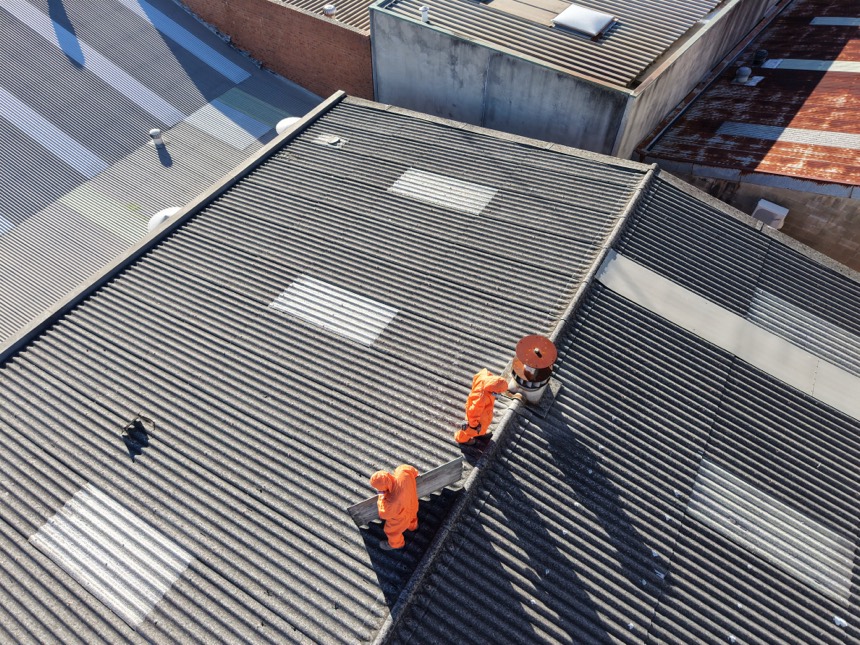 Licensed HazWreck crew in PPE removing a corrugated asbestos cement roof, viewed from above